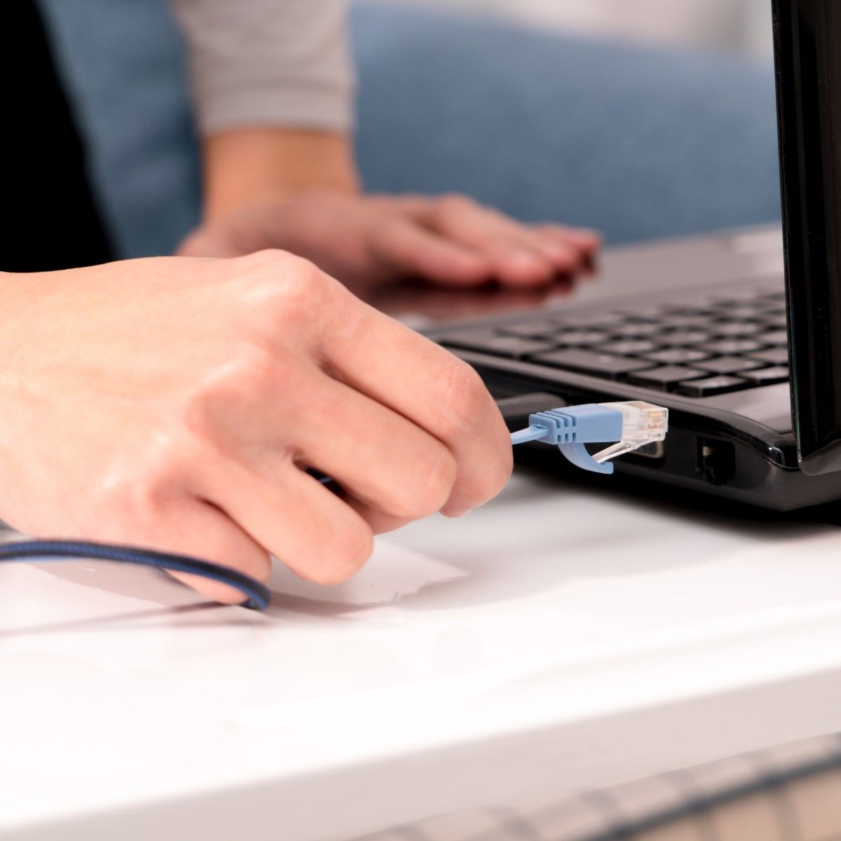 A close-up of a white woman&#x27;s right hand plugging a blue Ethernet cable into the side of her black laptop.