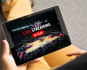 A person sitting on the ground holding a tablet in one hand and watching a livestream of a sports broadcast. The screen says &quot;Live Streaming&quot; and shows a basketball court.
