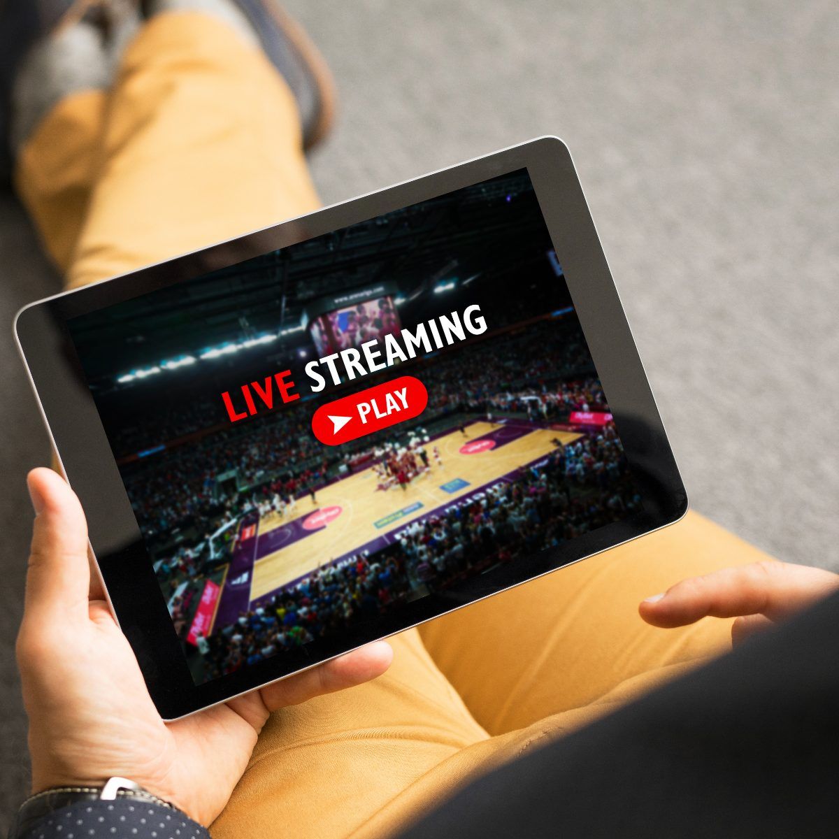 A person sitting on the ground holding a tablet in one hand and watching a livestream of a sports broadcast. The screen says &quot;Live Streaming&quot; and shows a basketball court.