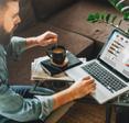 Man stirs cup of coffee that&#x27;s on a table on top of pile of books and next to mac laptop with graphics on the screen