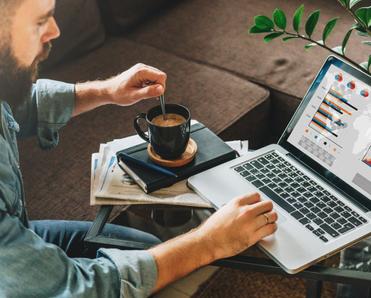 Man stirs cup of coffee that&#x27;s on a table on top of pile of books and next to mac laptop with graphics on the screen