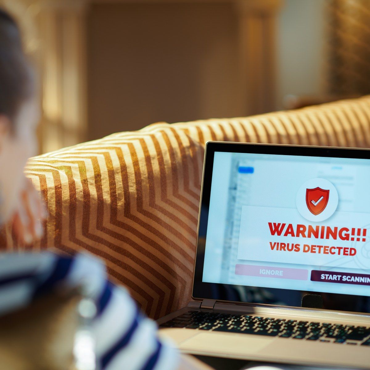 A woman sits on a yellow couch with a laptop in her lap, the screen says that a virus has been detected and asks if she wants to start a scan