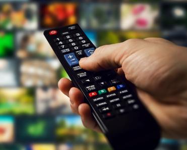 A hand holds a remote control pointed at a TV screen with multiple channels and shows displayed.