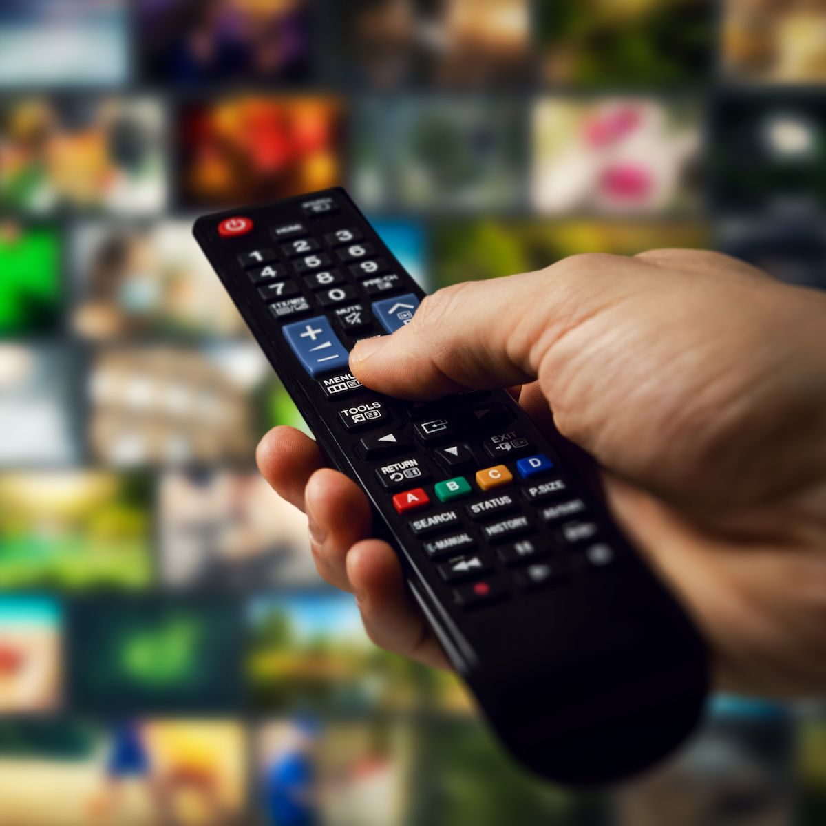 A hand holds a remote control pointed at a TV screen with multiple channels and shows displayed.