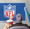 Person&#x27;s back as they wear American flag cap, popcorn and soda on one hand, and a giant foam hand in front of TV that shows NFL 