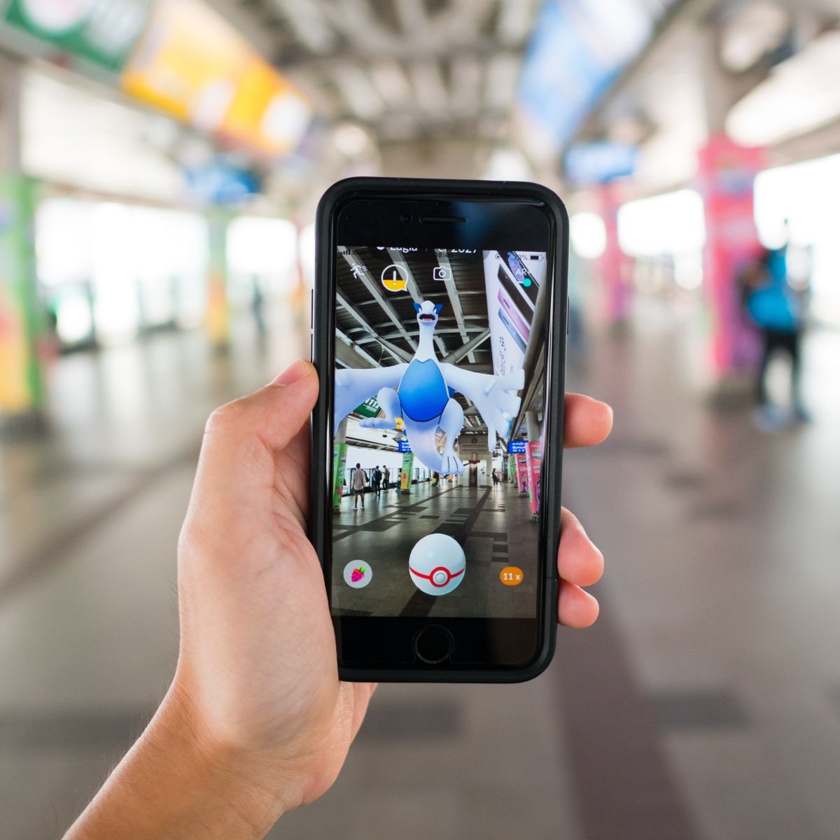 Open public space blurred in the back with hand holding phone screen shows app with game Pokemon GO