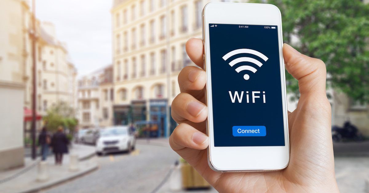 A hand holding a smartphone on a busy city street. The phone screen shows an oversized Wi-Fi signal.