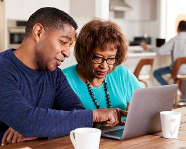 A middle-aged black man helps his older mother use social media safety tips on their home computer.