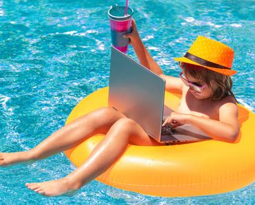 A little boy sits on a yellow floatation device in a swimming pool while using a laptop.