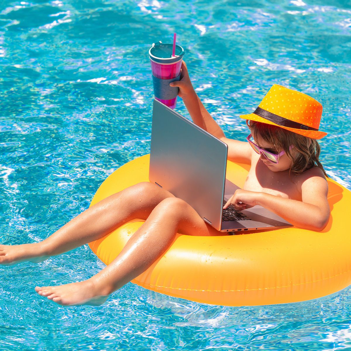 A little boy sits on a yellow floatation device in a swimming pool while using a laptop.