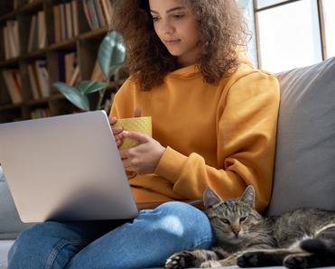 A young woman sits on a sofa with her pet cat and watches a TV show on her laptop. VPNs can help you unblock foreign streaming content.