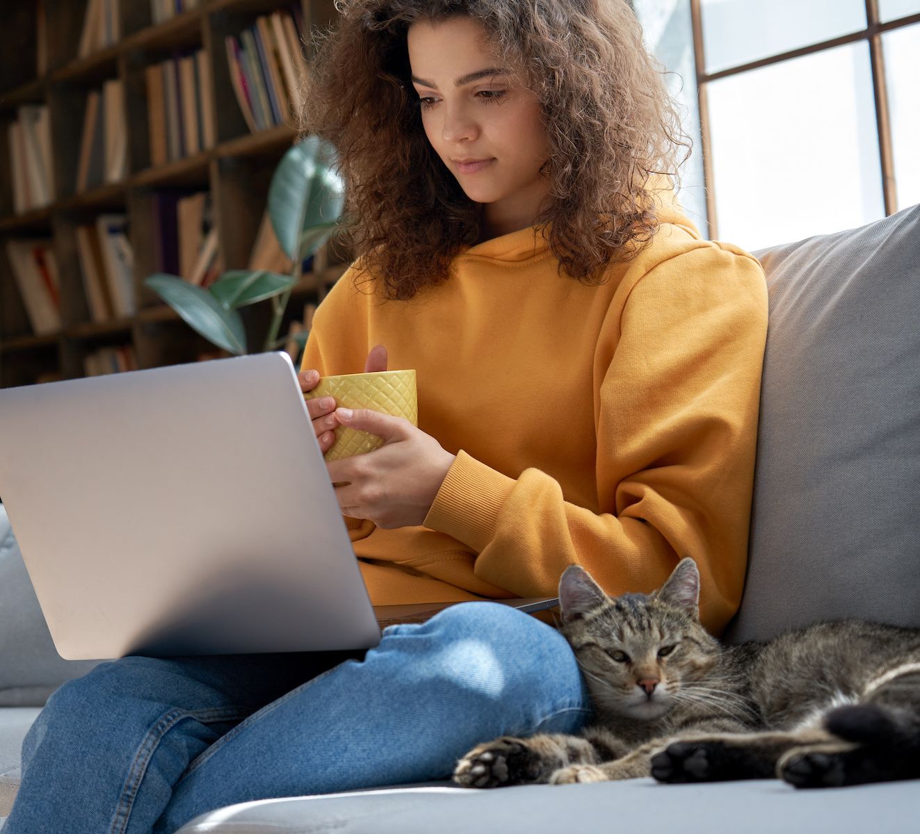 A young woman sits on a sofa with her pet cat and watches a TV show on her laptop. VPNs can help you unblock foreign streaming content.