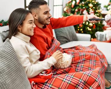 Young couple is sitting on the couch by their Christmas tree while the man is turning on the TV.