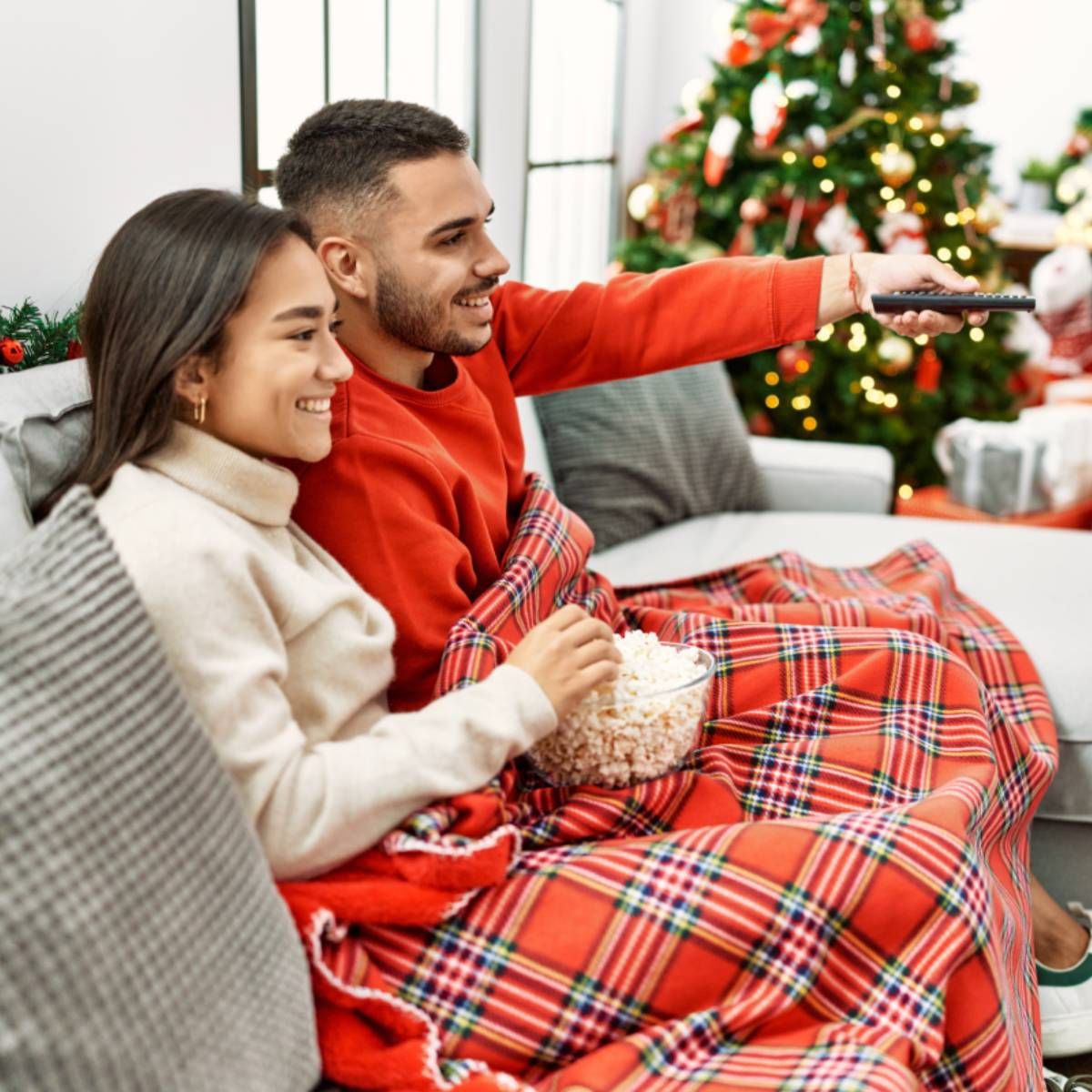 Young couple is sitting on the couch by their Christmas tree while the man is turning on the TV.