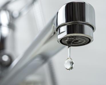 A close-up photo of a stainless steel kitchen sink faucet with a single drop of water leaking out of it.