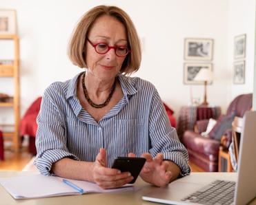A senior woman with light brown hair and red glasses sits at a desk and checks her cell phone. Her laptop is open beside her.