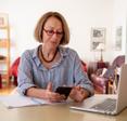 A senior woman with light brown hair and red glasses sits at a desk and checks her cell phone. Her laptop is open beside her.