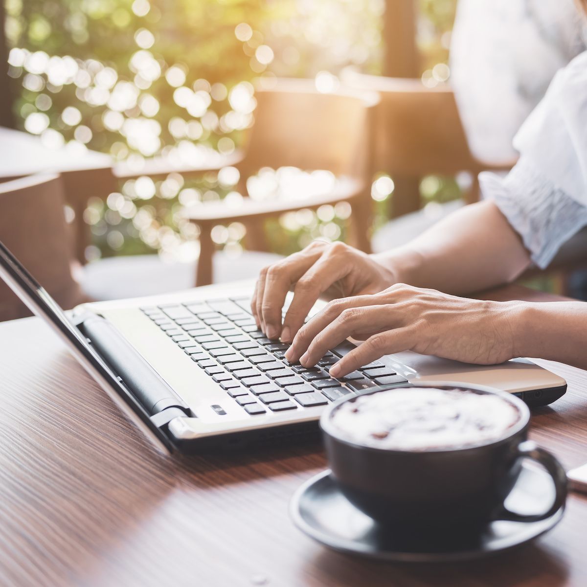 A woman&#x27;s hands rest on a laptop keyboard while she sits in a coffee shop. Using unsecure public Wi-Fi could put you at risk for a man-in-the-middle attack.