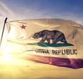 The state flag of California flutters against a blue sky and bright sunrise. The flag features a bear walking on green grass with a red star above it and the words &quot;California Republic&quot; below it.