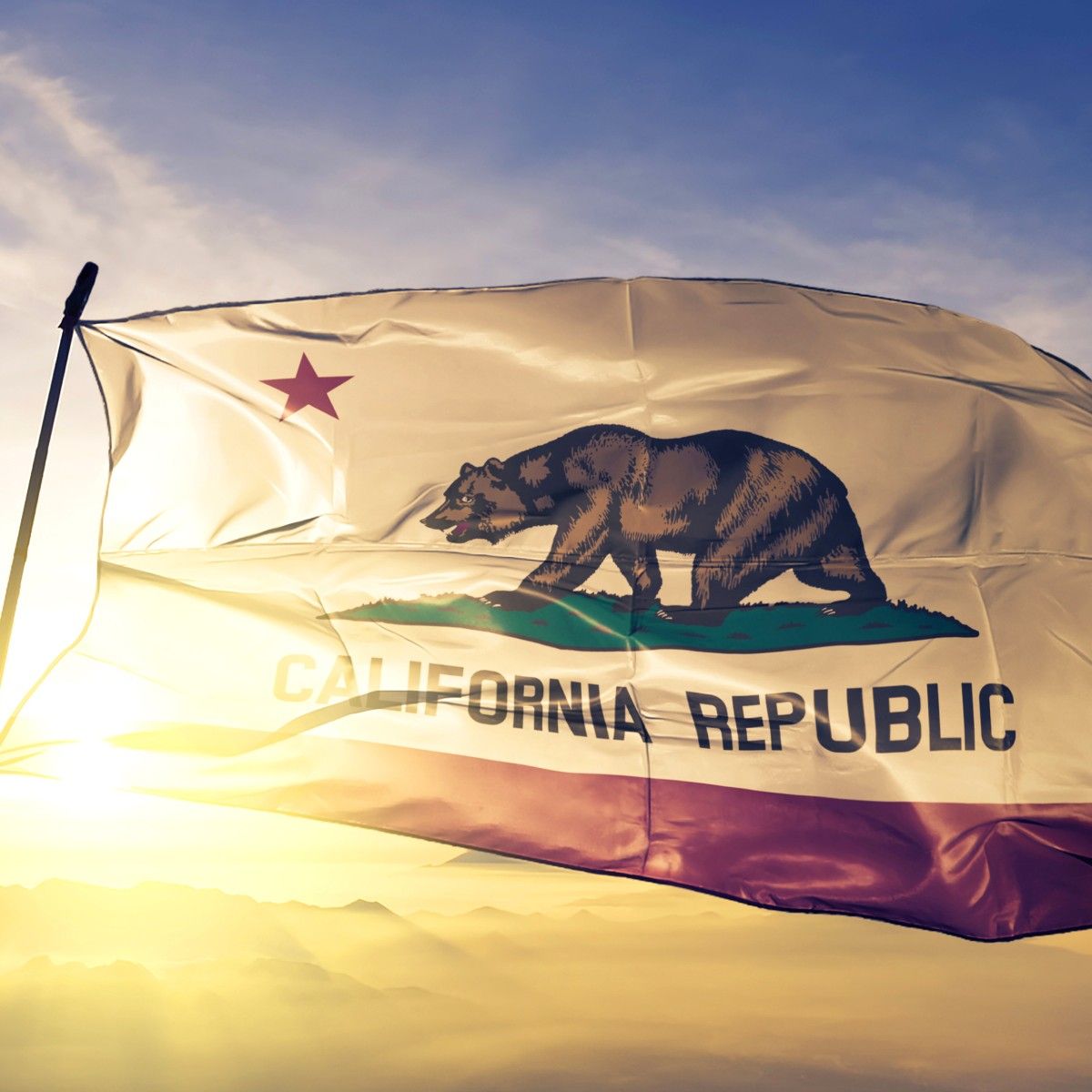 The state flag of California flutters against a blue sky and bright sunrise. The flag features a bear walking on green grass with a red star above it and the words &quot;California Republic&quot; below it.