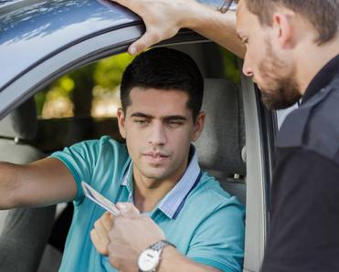 A young man showing his driver&#x27;s license and vehicle documentation to a cop from his car.