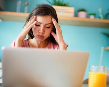 Woman with hands on her head looking frustrated while in front of laptop, glass of orange juice on the side and shelf on the back wall