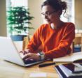 A dark-haired woman wearing glasses and an orange sweater types on her laptop.