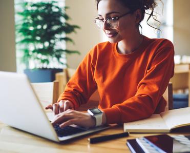 A dark-haired woman wearing glasses and an orange sweater types on her laptop.