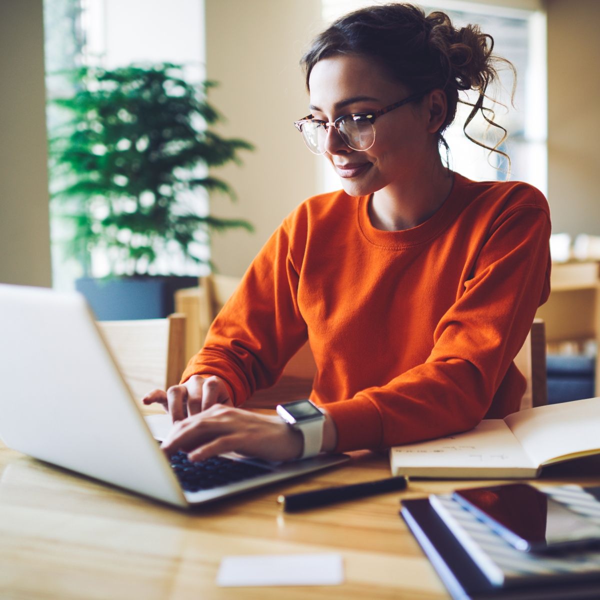 A dark-haired woman wearing glasses and an orange sweater types on her laptop.