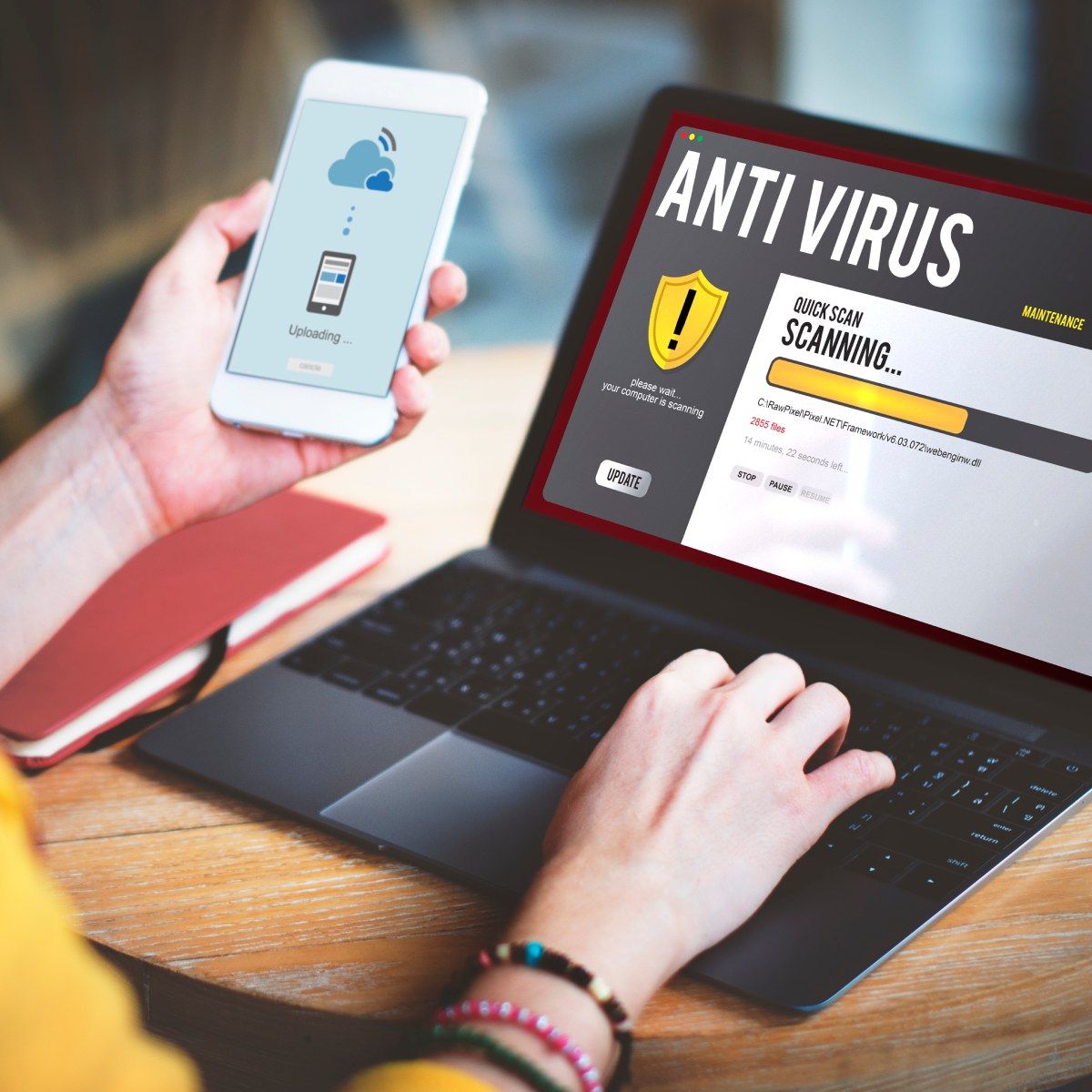 A woman&#x27;s hands hold her cell phone and type on her laptop while an antivirus software scan is on the screen