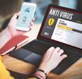 A woman&#x27;s hands hold her cell phone and type on her laptop while an antivirus software scan is on the screen