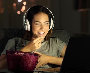 Woman smiling watching show on laptop with headphones on while eating popcorn