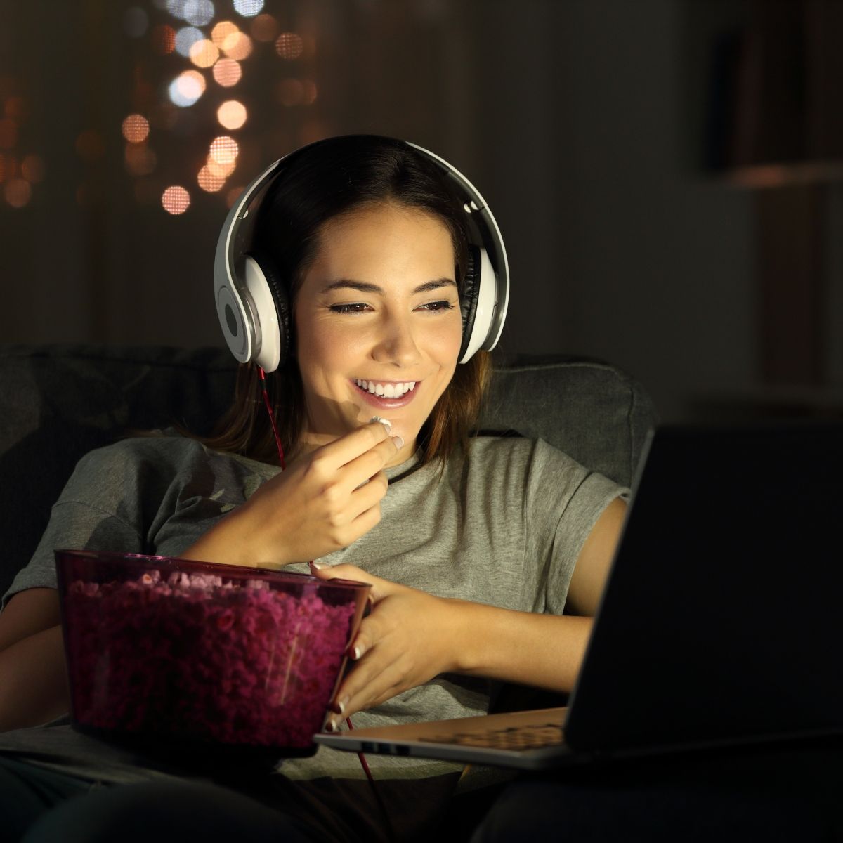 Woman smiling watching show on laptop with headphones on while eating popcorn