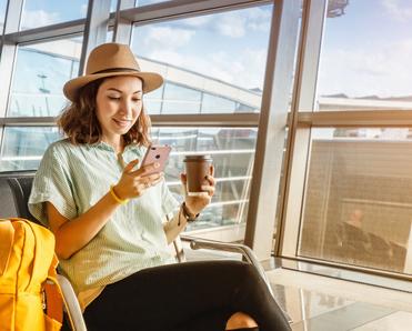 A young Asian woman sits next to her airport gate while using a cell phone and drinking coffee.