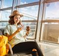 A young Asian woman sits next to her airport gate while using a cell phone and drinking coffee.