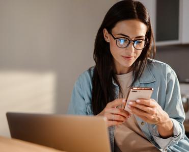 Dark-haired woman with glasses is looking down at her phone with a pleasant expression. 