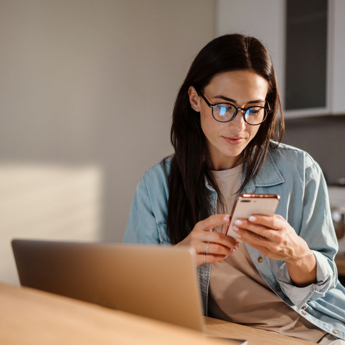 Dark-haired woman with glasses is looking down at her phone with a pleasant expression. 