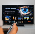 A hand holds a TV remote in front of a plasma TV screen showing the Netflix library and a Black Mirror feature