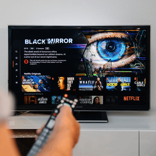 A hand holds a TV remote in front of a plasma TV screen showing the Netflix library and a Black Mirror feature