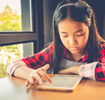 An Asian girl plays with a tablet computer at a table in her home next to a window.