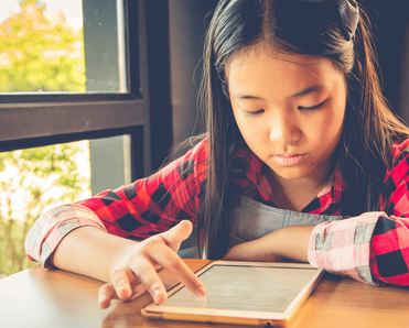 An Asian girl plays with a tablet computer at a table in her home next to a window.