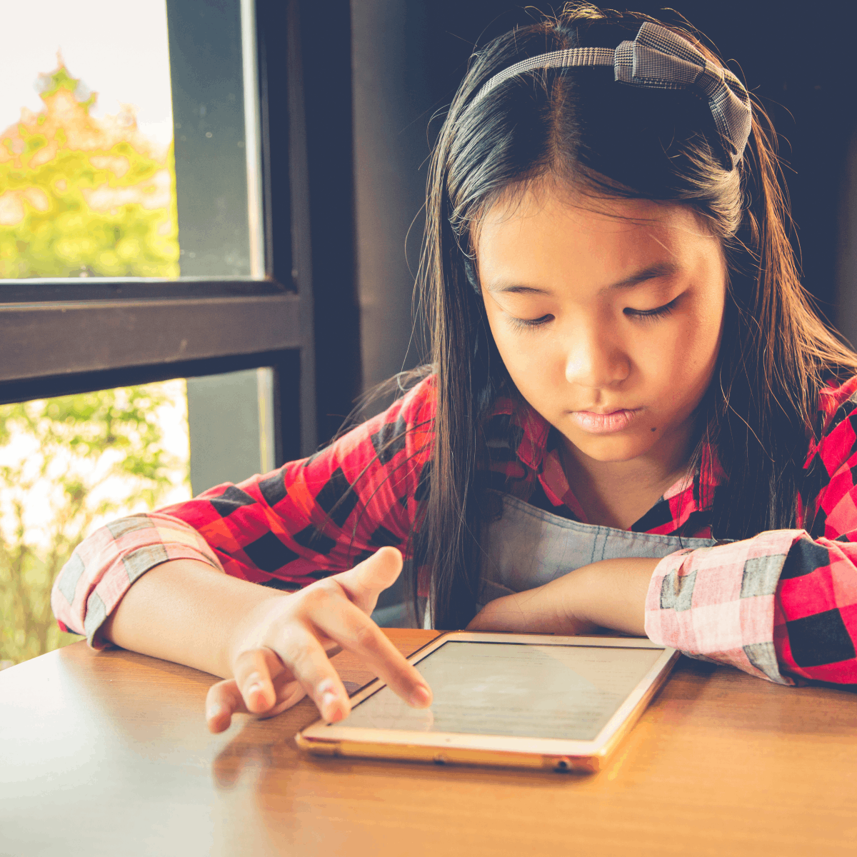 An Asian girl plays with a tablet computer at a table in her home next to a window.